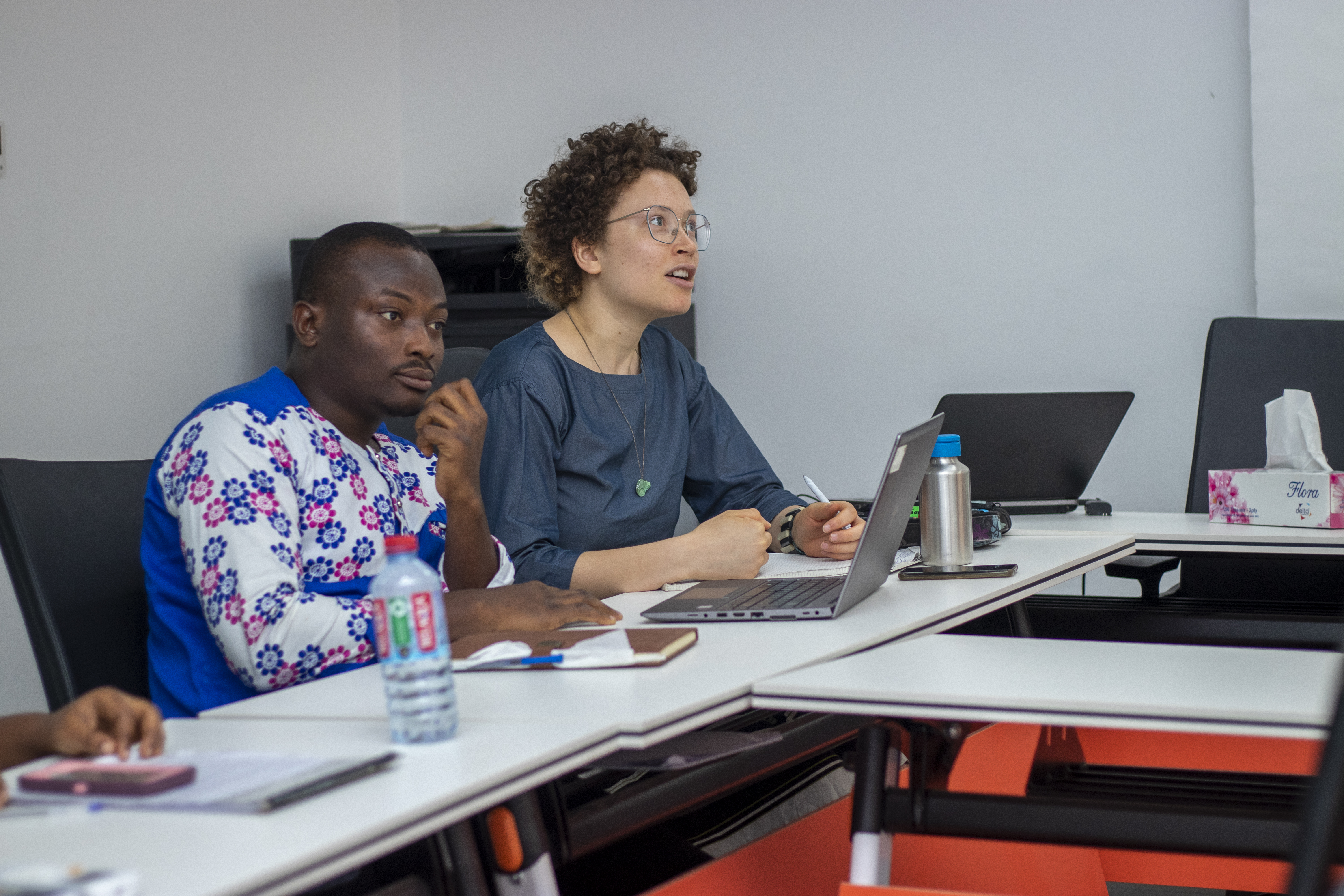Two professionals at a desk in a bright room, focused on a speaker; laptop and notes suggest a workshop or seminar.